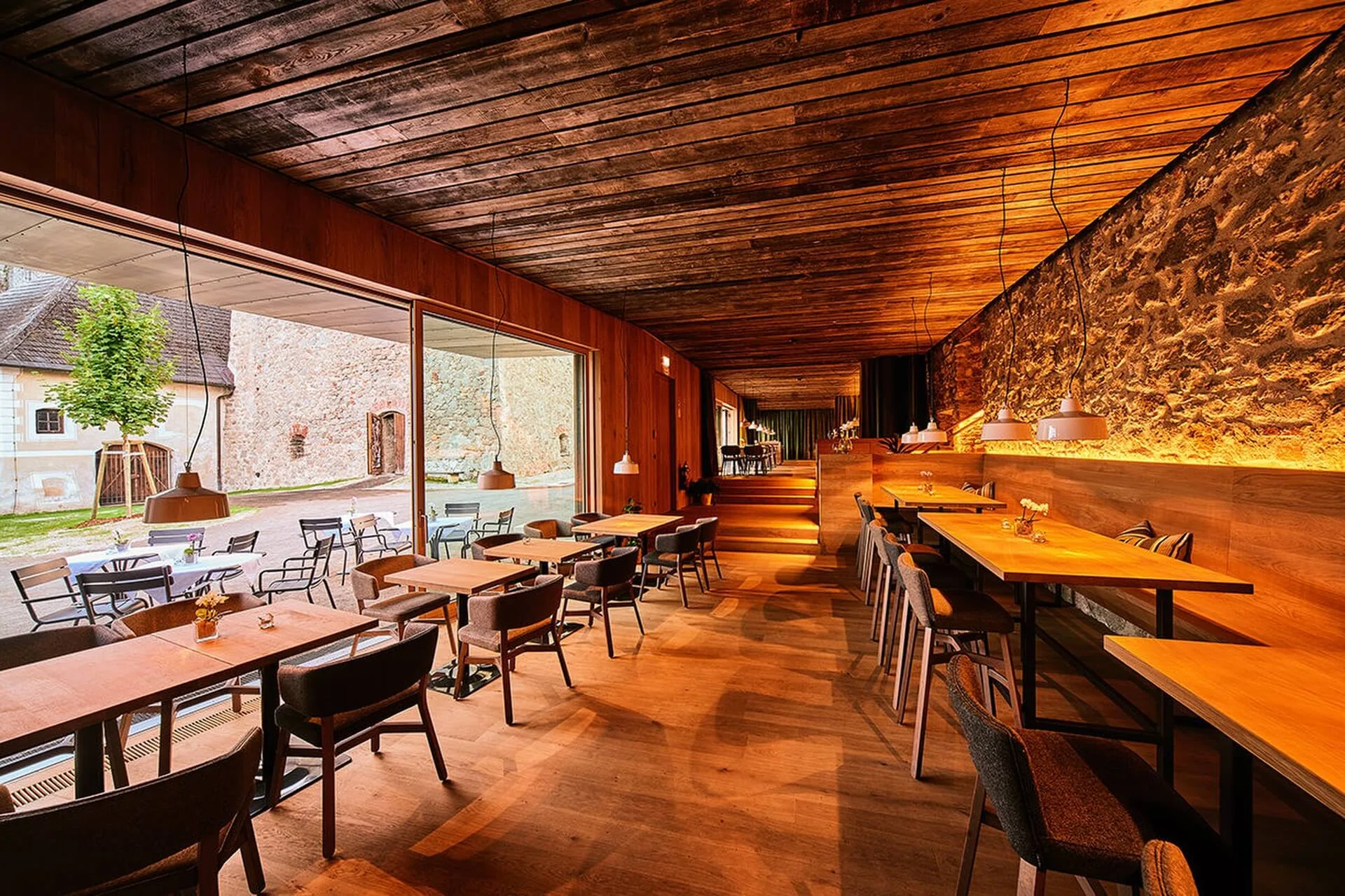 Modernes Restaurant mit Holzböden, Decken und Steinwänden. Große Fenster bieten einen Blick auf einen Außenbereich mit Tischen und Stühlen. Die warme Beleuchtung schafft eine gemütliche Atmosphäre.