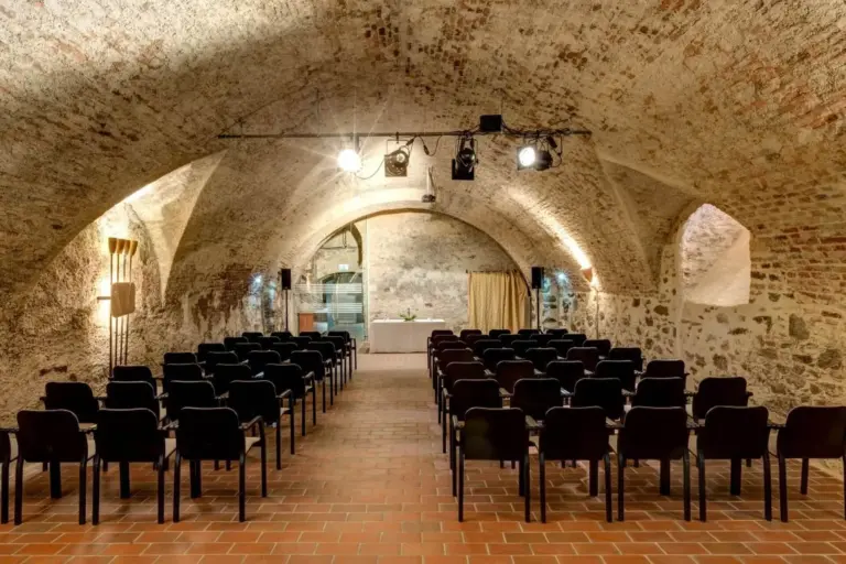 Ein gewölbter Steinsaal mit schwarzen Stuhlreihen vor einer kleinen Bühne, die von Deckenstrahlern beleuchtet wird und dem Raum eine rustikale und historische Atmosphäre verleiht.