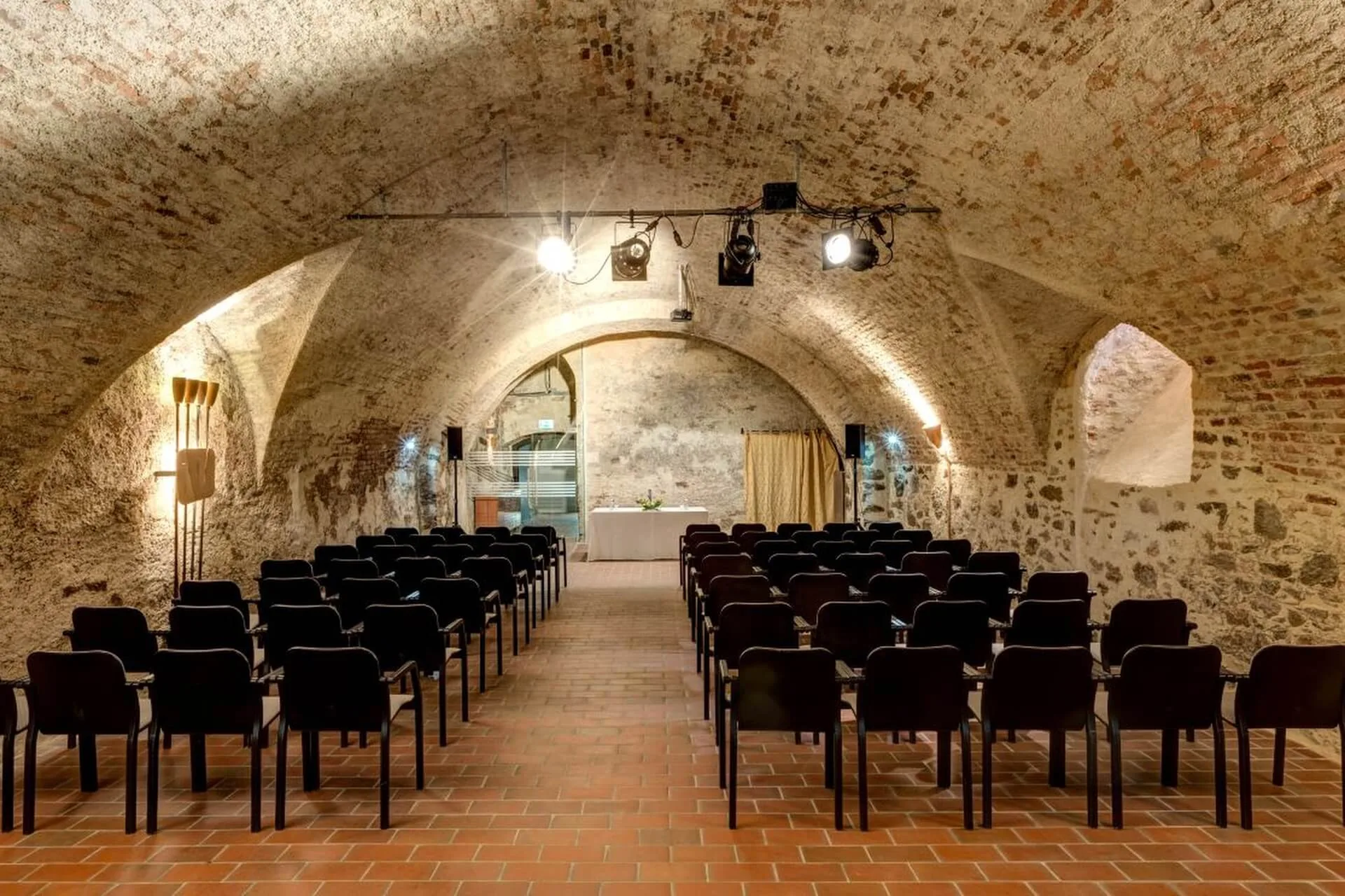 Ein gewölbter Steinsaal mit schwarzen Stuhlreihen vor einer kleinen Bühne, die von Deckenstrahlern beleuchtet wird und dem Raum eine rustikale und historische Atmosphäre verleiht.