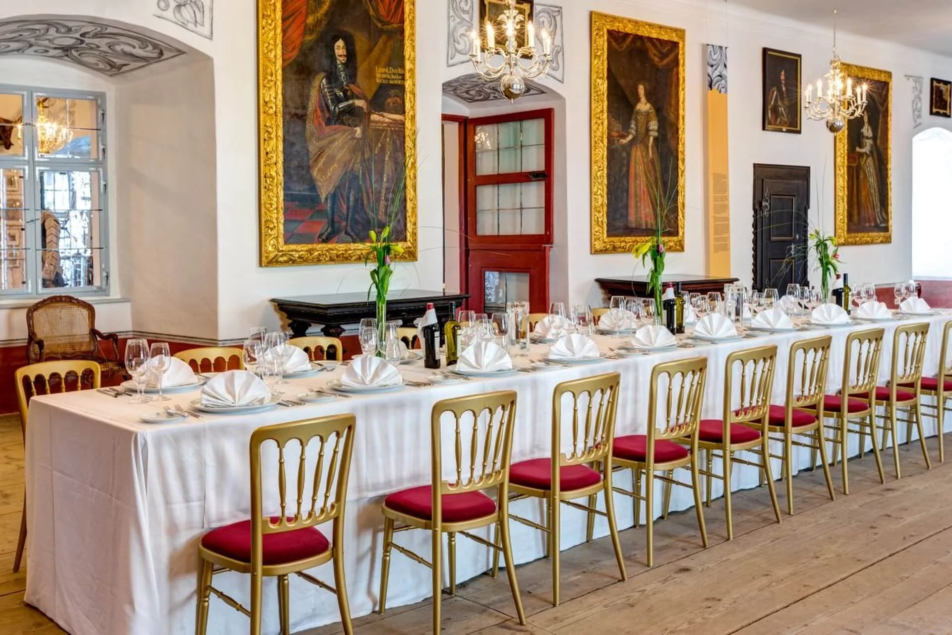 Ein langer, eleganter Esstisch, gedeckt für ein formelles Essen mit goldenen Stühlen, gefalteten Servietten, Gläsern und weißen Tischtüchern in einem großen Raum mit großen Porträts und verzierten Kronleuchtern.