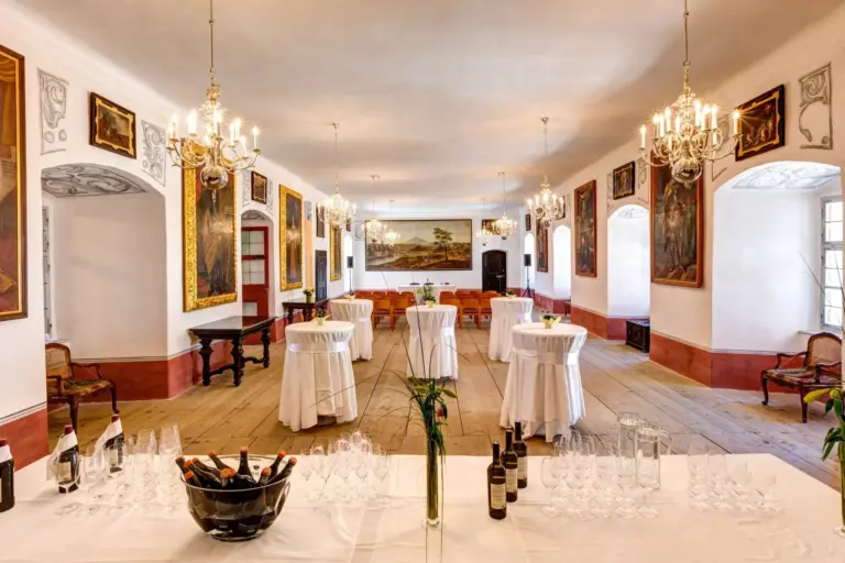 Eleganter Bankettsaal mit Kronleuchtern, runden, mit weißen Tüchern bedeckten Tischen, Weinflaschen und Gläsern, die auf einem Tisch im Vordergrund angeordnet sind. Die Wände sind mit großen Gemälden und verschnörkelten Leisten verziert.