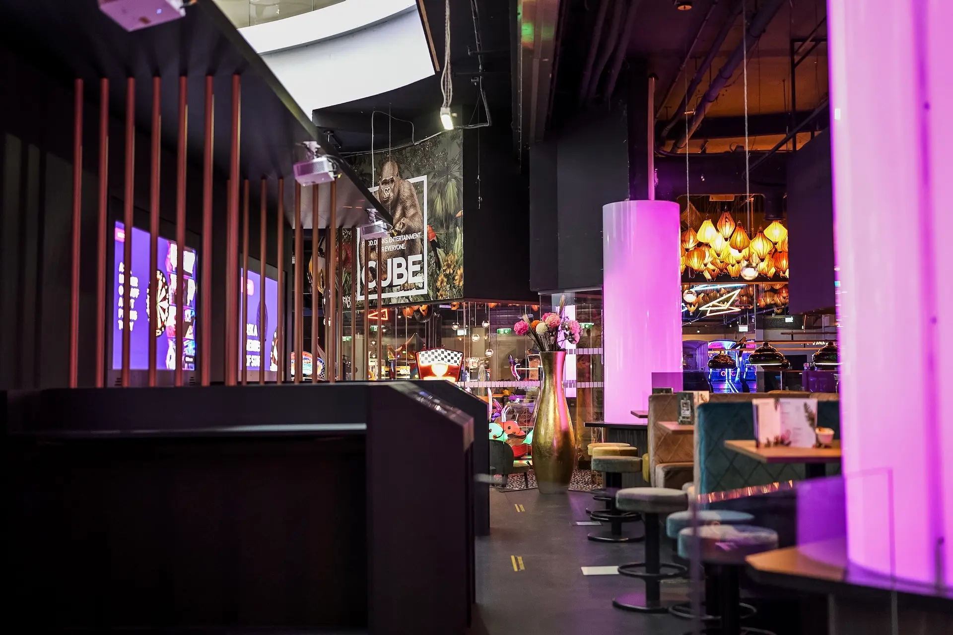 Eine moderne, stilvolle Bar oder Lounge mit violetter Beleuchtung, Plüschsitzen, hohen Vasen mit Blumen und dekorativen Hängelampen. An der Wand im Hintergrund ist ein Filmplakat zu sehen.