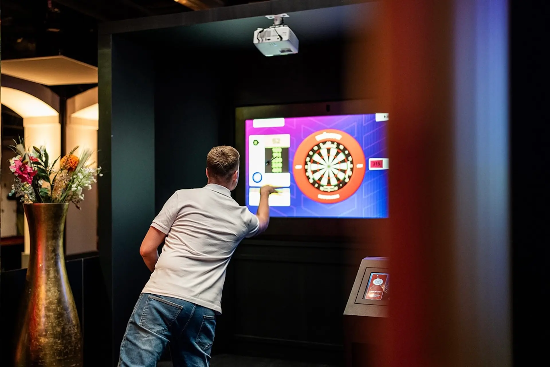 Ein Mann in weißem Hemd und Jeans wirft einen Dartpfeil auf eine digitale Dartscheibe, die auf einem großen Bildschirm in einer modernen Inneneinrichtung angezeigt wird. Auf der linken Seite steht eine hohe Vase mit bunten Blumen.