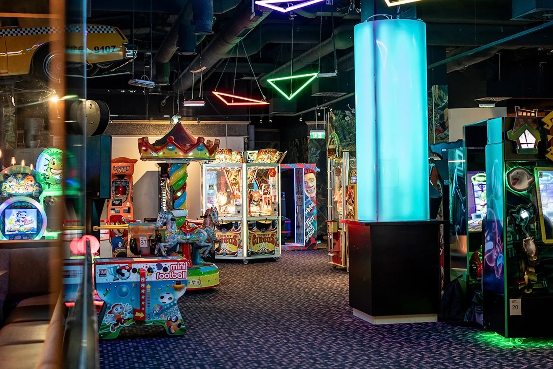 Eine bunte Spielhalle mit verschiedenen Spielautomaten, hellen Neonlichtern und einer leuchtenden Säule in der Mitte. Der Boden ist mit Teppich ausgelegt, und überall im Raum gibt es Fahrgeschäfte und blinkende Spielbildschirme.