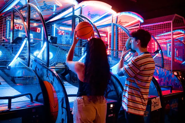 Zwei Personen spielen ein Arcade-Basketballspiel unter hellen Neonlichtern. Eine Person bereitet sich darauf vor, einen Basketball zu werfen, während die andere einen Ball hält und zusieht. Die Szene ist lebendig und bunt.