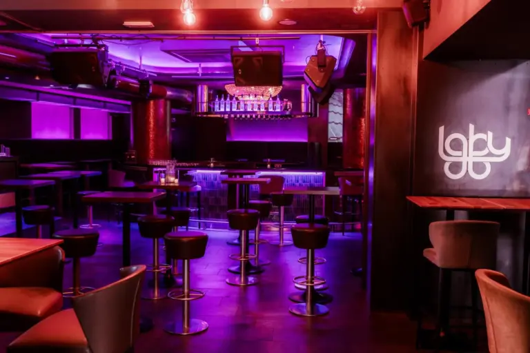 Ein moderner Nachtclub mit lila und pinkfarbener Beleuchtung, mit Stehtischen, Barhockern, einer beleuchteten Bar und einem Laby-Logo an der Wand. Der Club ist leer und hat eine stilvolle, einladende Atmosphäre.