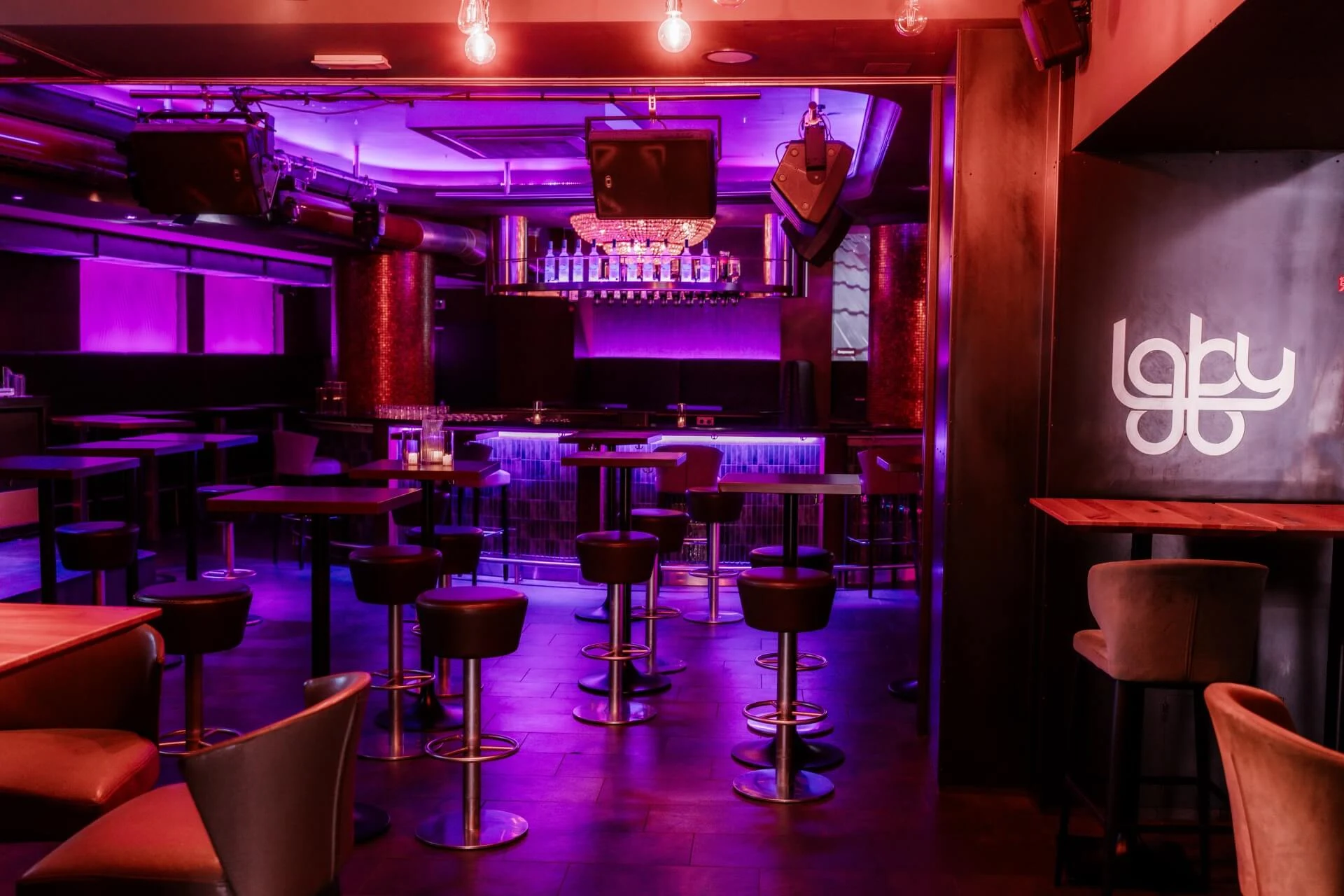 Ein moderner Nachtclub mit lila und pinkfarbener Beleuchtung, mit Stehtischen, Barhockern, einer beleuchteten Bar und einem Laby-Logo an der Wand. Der Club ist leer und hat eine stilvolle, einladende Atmosphäre.