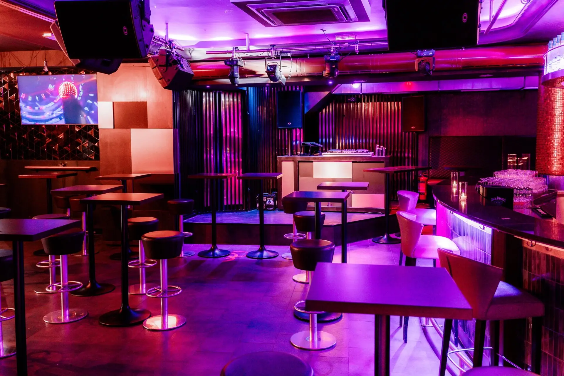 Ein moderner Nachtclub mit lila und pinkfarbener Beleuchtung, hohen Tischen und Hockern, einer DJ-Kabine, an der Wand befestigten Lautsprechern und einem Fernseher, der ein buntes Bild zeigt. Der Raum ist leer, und die Atmosphäre ist lebendig und stilvoll.