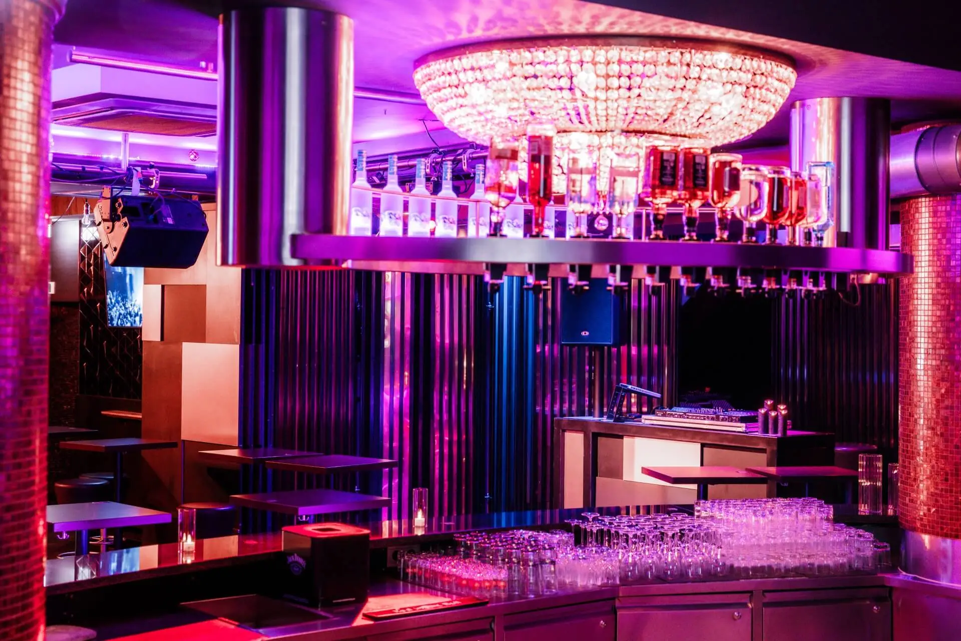 Ein moderner Nachtclub mit lila und rosa Beleuchtung, einem großen Kronleuchter über einer Bar mit Gläsern und Flaschen, leeren Tischen, Barhockern und einem DJ-Pult im Hintergrund.