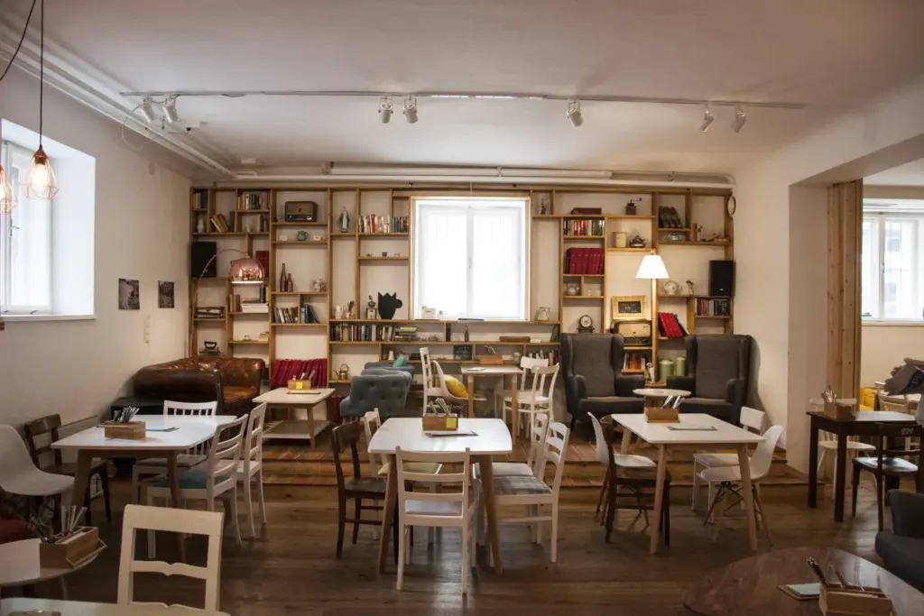 Eine gemütliche Café-Caspar-Einrichtung mit Holzböden, uneinheitlichen Tischen und Stühlen, Bücherregalen an der Rückwand, bequemen Sesseln und sanfter Beleuchtung durch Fenster und Lampen.