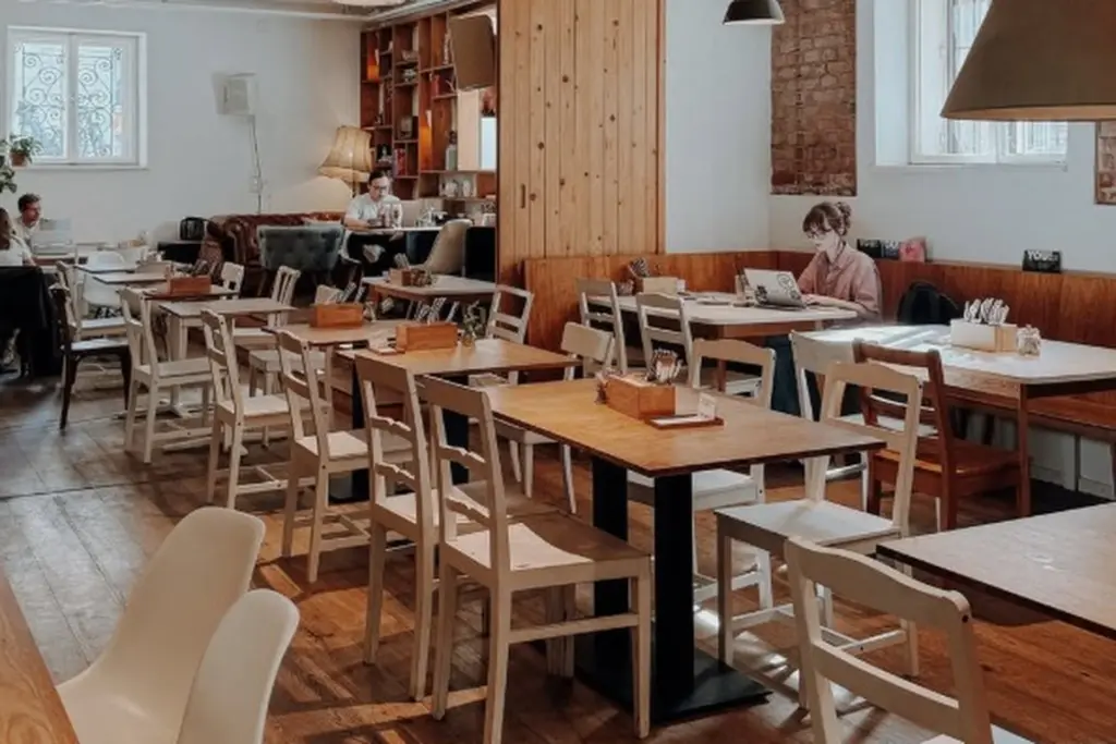 Ein gemütliches Café mit Holztischen und -stühlen, ein paar Leuten, die an Laptops arbeiten, und natürlichem Licht, das durch große Fenster fällt. Der Raum hat eine warme Beleuchtung, Backsteinakzente und Bücherregale im Hintergrund.