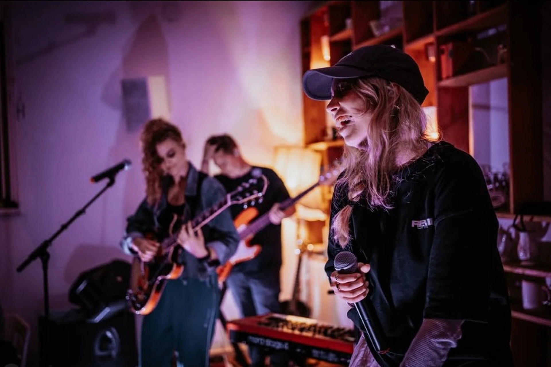 Eine kleine Band spielt in einem Innenraum des Café Caspar. Eine Frau mit Mütze singt in ein Mikrofon, während eine andere eine E-Gitarre spielt und eine dritte Person im Hintergrund den Bass bedient. Die warme Beleuchtung schafft eine gemütliche, intime Atmosphäre.