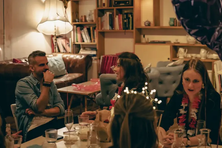 Vier Personen sitzen um einen Tisch im gemütlichen, warm beleuchteten Café Caspar, unterhalten sich und genießen Getränke. Bücherregale, Sofas und sanfte Lampen schaffen im Hintergrund eine entspannte, einladende Atmosphäre.
