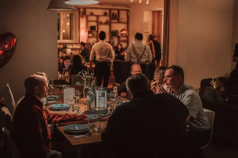 Eine Gruppe von Menschen sitzt um einen Tisch im warm beleuchteten Café Caspar und unterhält sich. Im Hintergrund sind weitere Gäste und das Restaurantpersonal zu sehen, die eine gemütliche, lebhafte Atmosphäre schaffen.