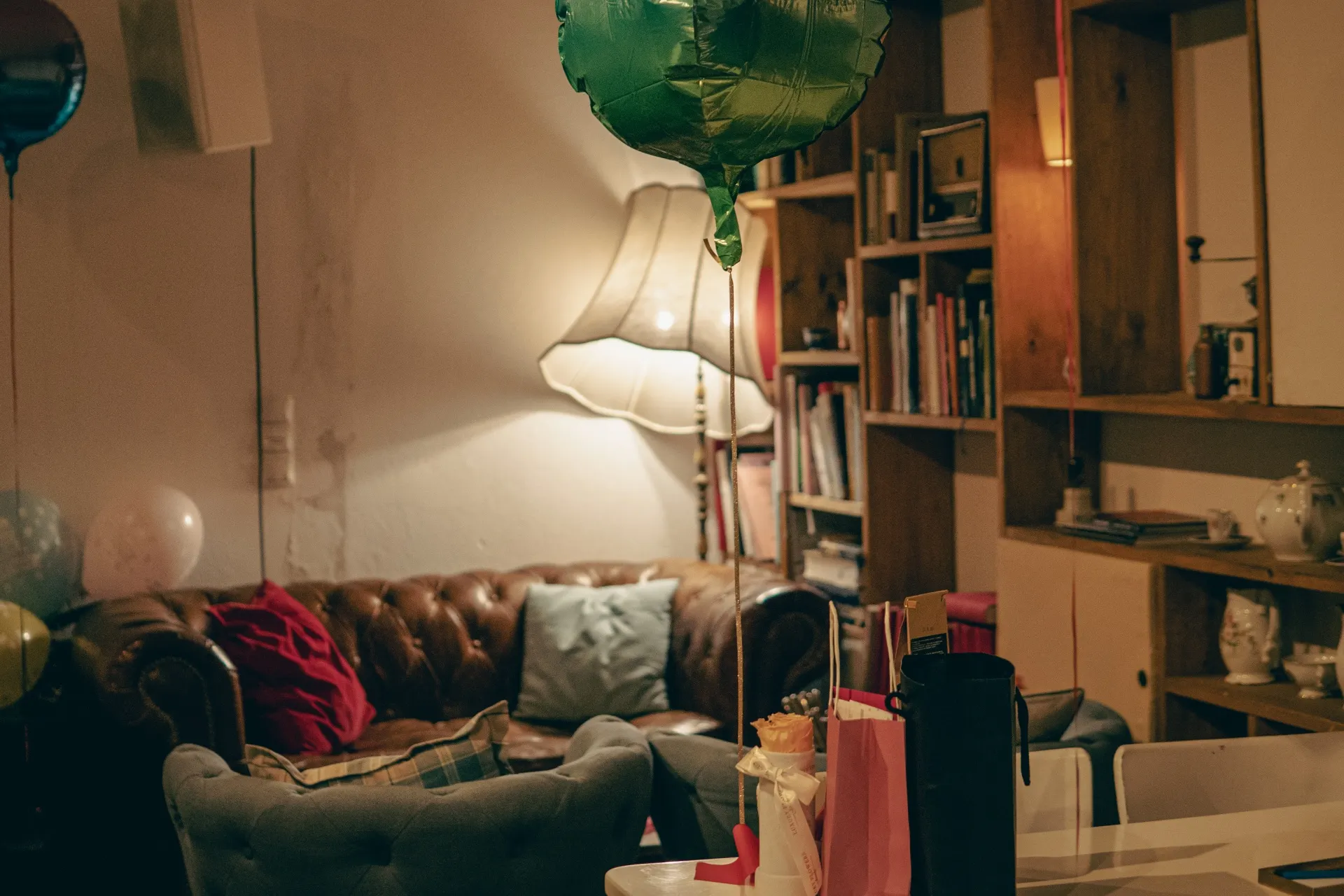 Ein gemütliches Wohnzimmer mit einem Ledersofa, einer Stehlampe, Bücherregalen und Geschenktüten auf einem Tisch - ganz wie die einladende Atmosphäre im Café Caspar. Warme Beleuchtung und ein grüner Ballon verleihen diesem entspannten Raum Charme.