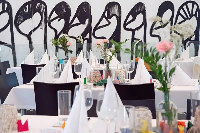 Ein modern gedeckter Restauranttisch mit weißen Servietten, Glaswaren und kleinen Blumenvasen; an der Wand im Hintergrund ist ein abstraktes schwarz-weißes Wandgemälde mit stilisierten Gesichtern zu sehen.