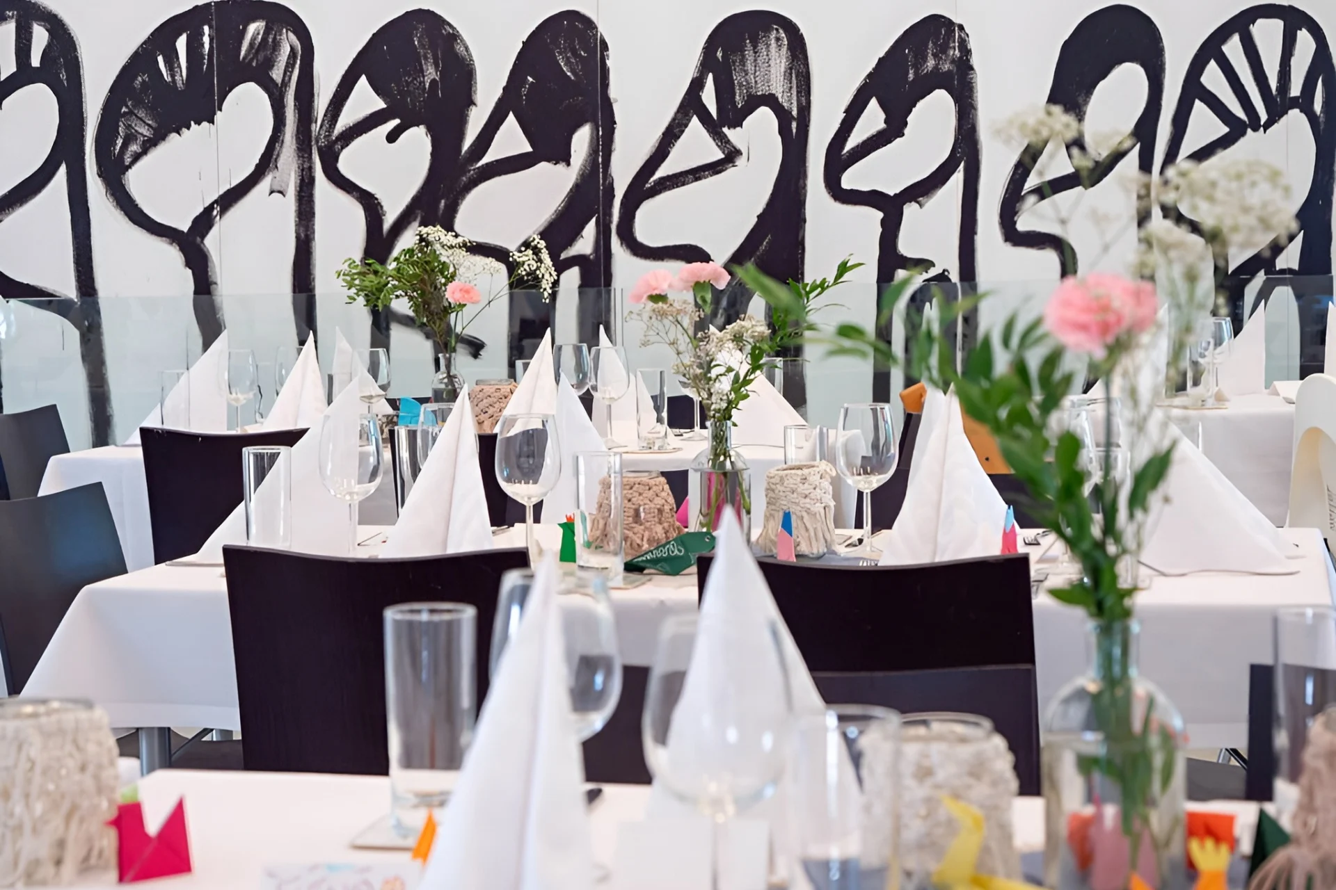 Ein modern gedeckter Restauranttisch mit weißen Servietten, Glaswaren und kleinen Blumenvasen; an der Wand im Hintergrund ist ein abstraktes schwarz-weißes Wandgemälde mit stilisierten Gesichtern zu sehen.