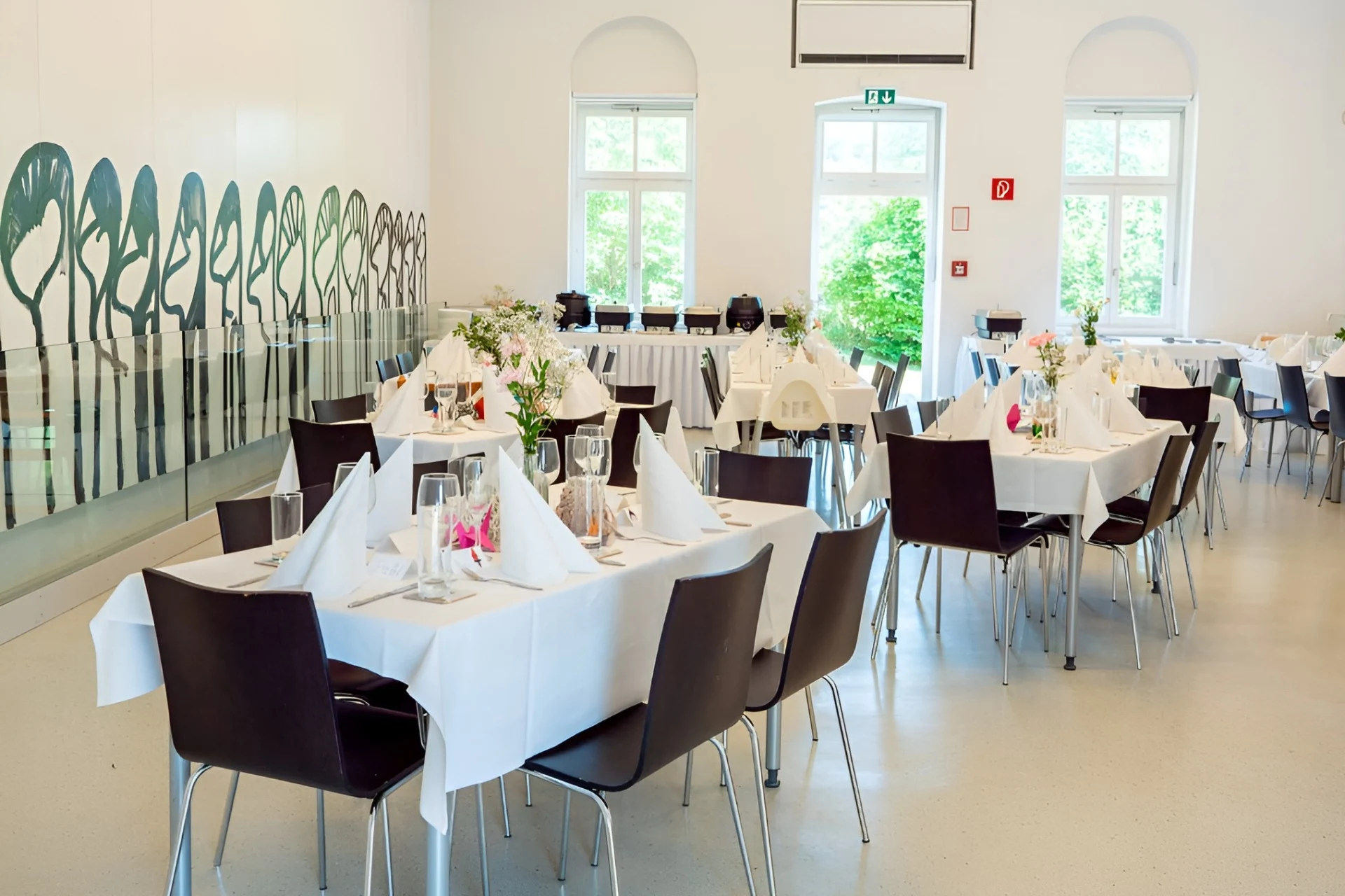 Ein heller Bankettsaal mit weißen Wänden und großen Fenstern, der für eine Veranstaltung eingerichtet ist. Die Tische sind mit weißen Tüchern, gefalteten Servietten, Gläsern und Blumensträußen eingedeckt. Dunkle Stühle umgeben jeden Tisch.