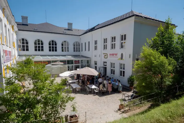 Eine Gruppe von Menschen versammelt sich an einem sonnigen Tag unter Sonnenschirmen im Innenhof eines weißen Gebäudes mit großen Fenstern. Einige Grünflächen und Bäume umgeben den Innenhof.