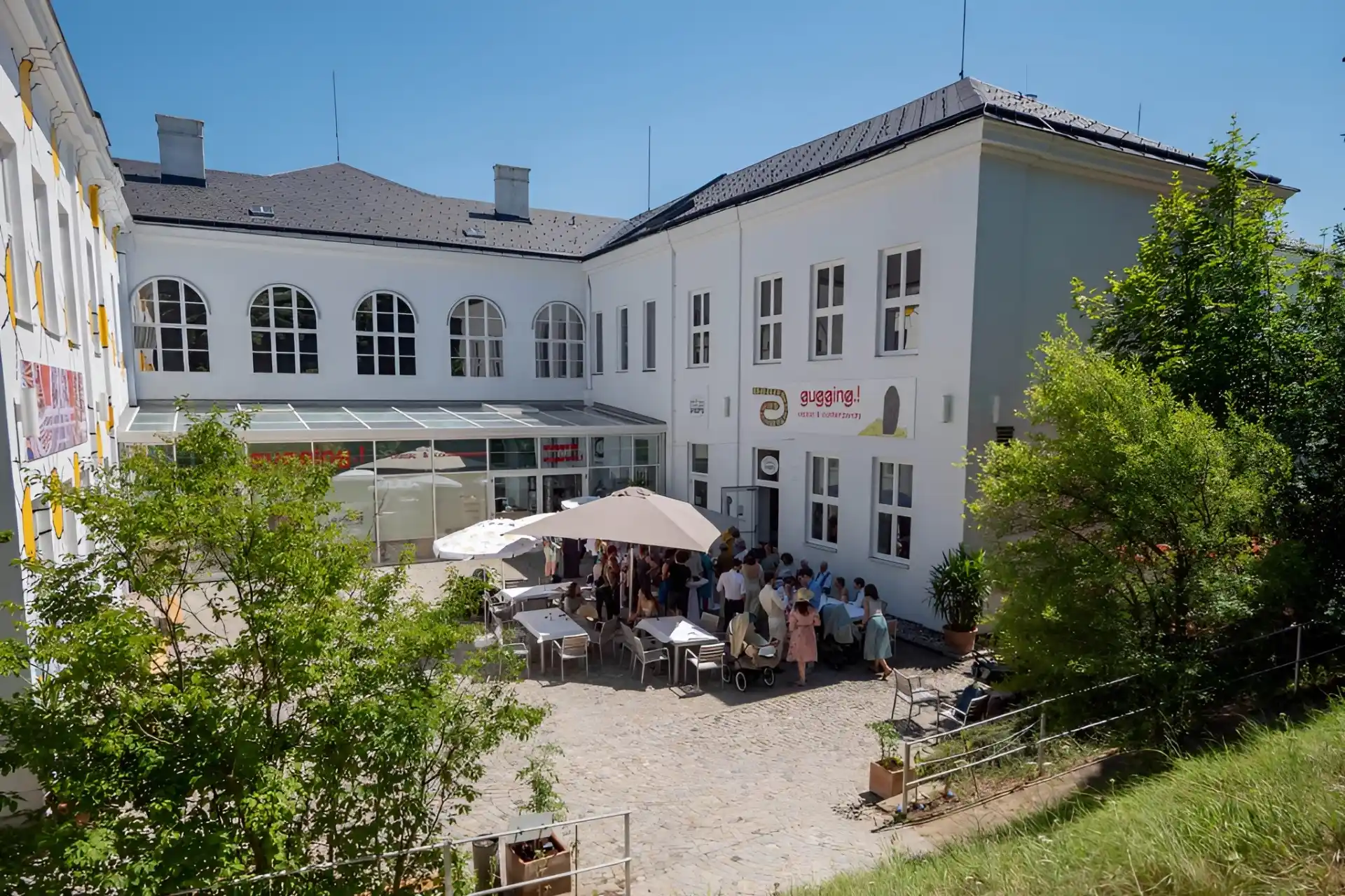 Eine Gruppe von Menschen sitzt an einem sonnigen Tag unter Sonnenschirmen an Tischen im Freien in einem Innenhof zwischen zwei weißen Gebäuden. Im Vordergrund sind Bäume und Grünpflanzen zu sehen; auf einem Schild an dem Gebäude steht "Bauernjause".