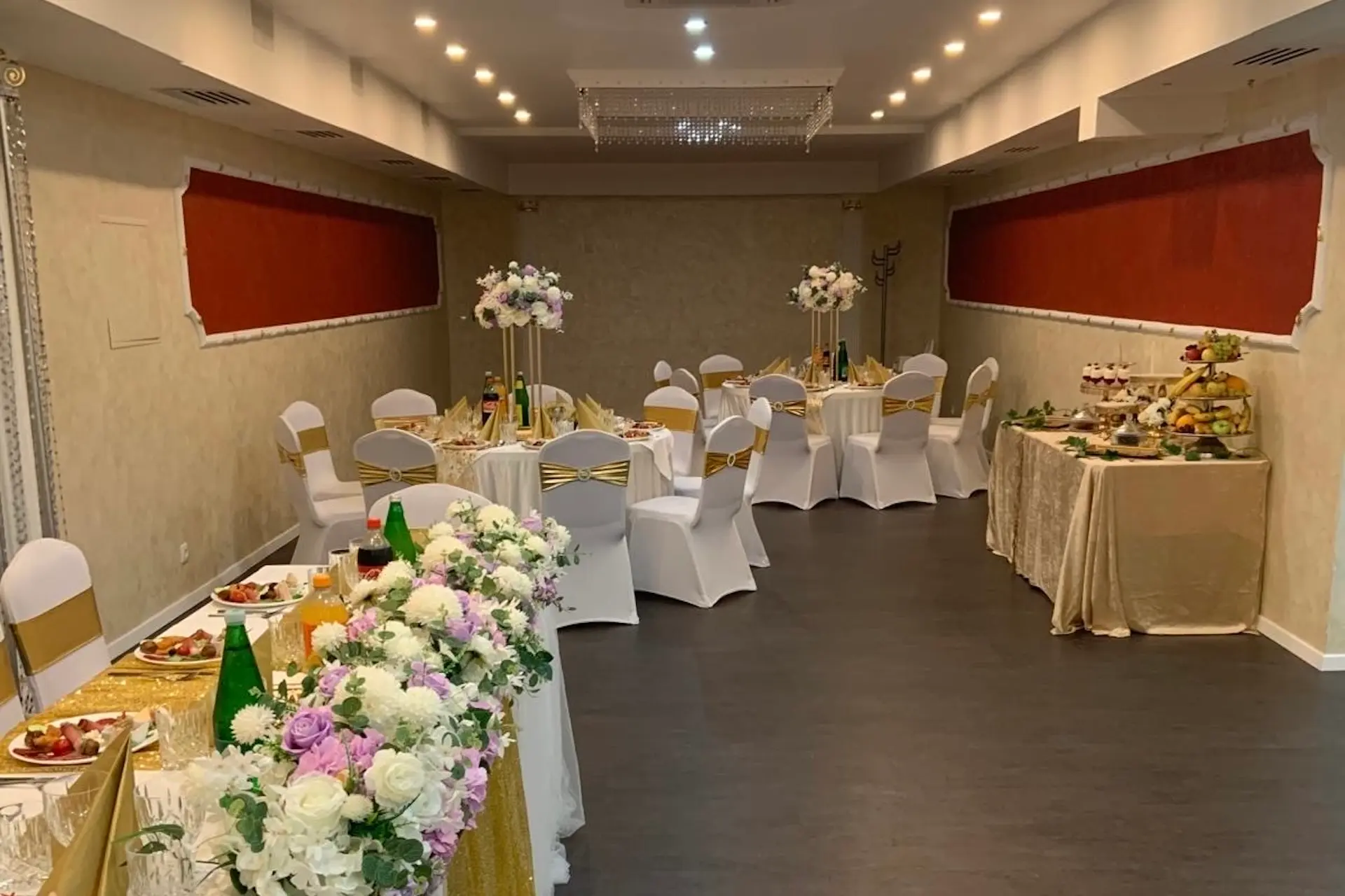 Eleganter Bankettsaal mit runden Tischen, die mit weißen Tüchern und goldenen Akzenten bedeckt und mit hohen Blumensträußen geschmückt sind, die für eine Veranstaltung arrangiert wurden. Ein langer Tisch mit Blumenarrangements und Speisen steht an der Stirnseite.