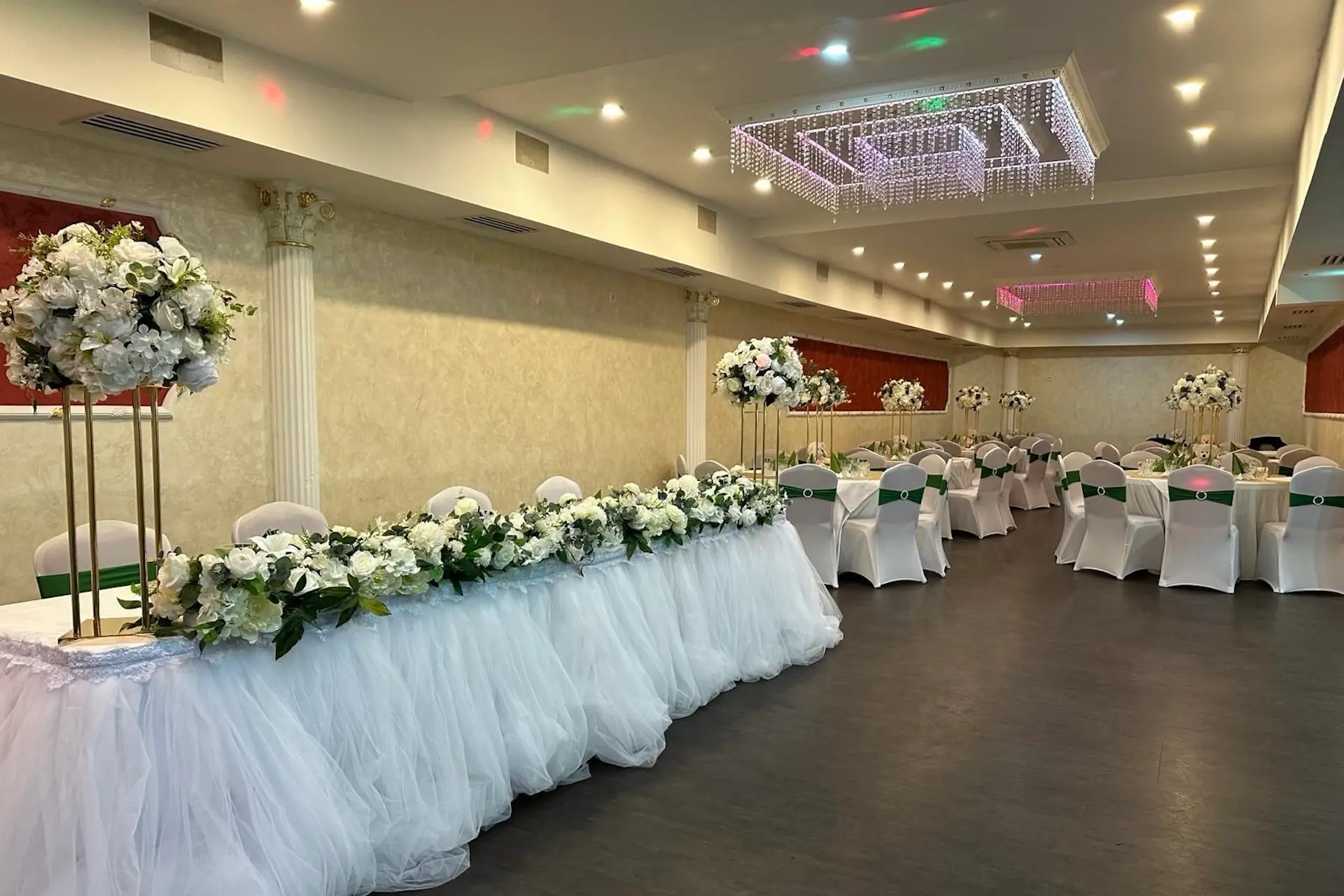 Ein dekorierter Bankettsaal mit weiß bezogenen Stühlen, grünen Schärpen und langen Tischen, die mit weißen Blumenarrangements und Tüll geschmückt sind. Elegante Kronleuchter hängen von der Decke und schaffen eine festliche Atmosphäre.