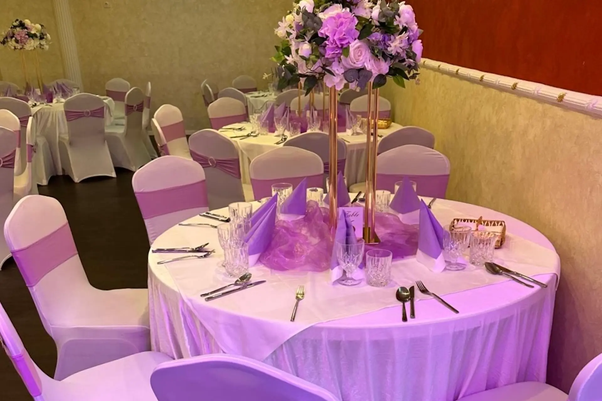 Ein runder Tisch für eine formelle Veranstaltung mit weißer Tischdecke, lila Servietten, Gläsern und Silberbesteck. Ein hoher Tafelaufsatz mit rosa und weißen Blumen steht in der Mitte; die Stühle haben weiße Bezüge mit lila Bändern.