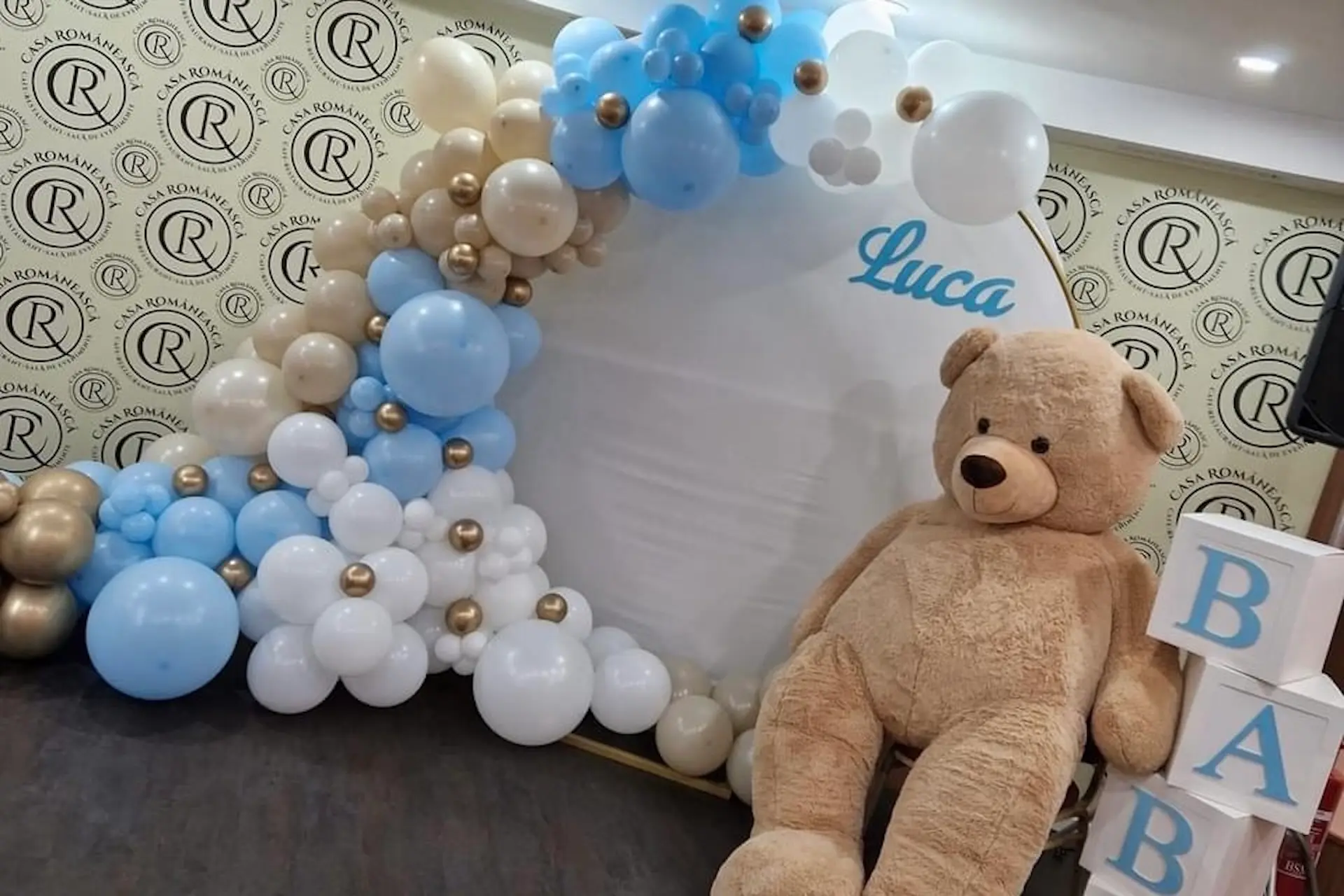Ein großer beiger Teddybär sitzt neben dekorativen Blöcken mit der Aufschrift BABY und einem weißen Zelt, das mit blauen, weißen und goldenen Luftballons geschmückt ist. Der Name Luca ist in blauer Schrift auf dem Zelt zu lesen.