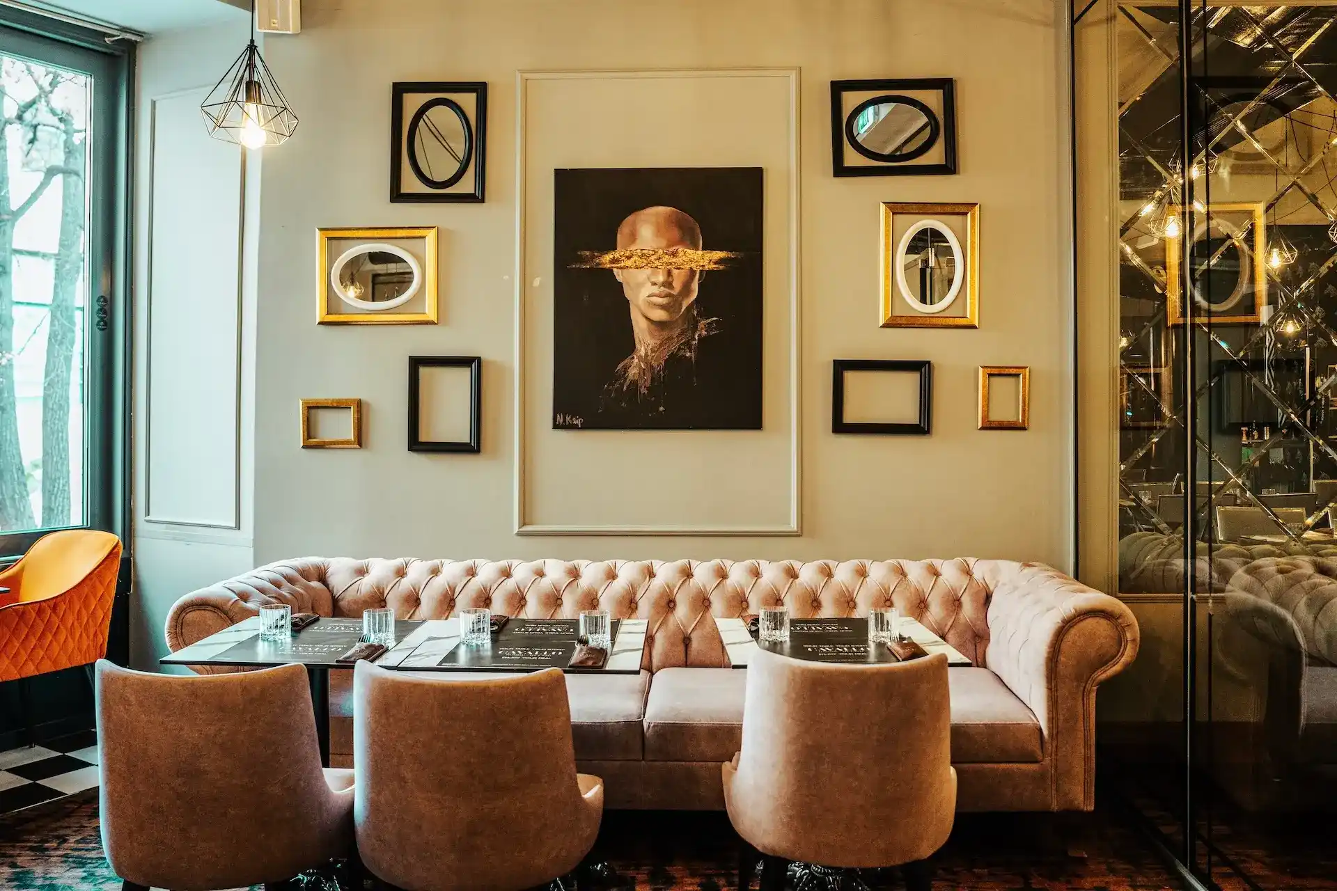Ein stilvolles Wohnzimmer mit einem getufteten beigen Sofa, Plüschsesseln und einem gläsernen Couchtisch. Die Wand dahinter zeigt leere goldene und schwarze Rahmen um das Porträt einer Person mit goldenen Schlieren über den Augen.