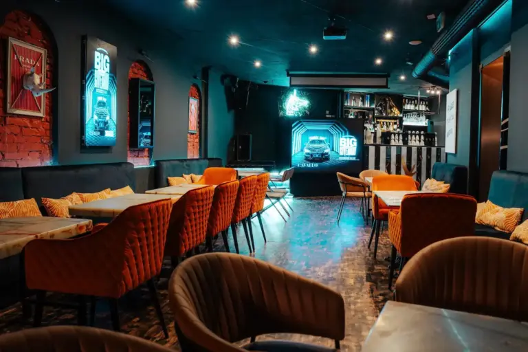 Cavallo Wien Eine moderne Lounge-Bar mit orangefarbenen und braunen Stühlen, kleinen Tischen, Leuchtreklamen, Postern und einem gut beleuchteten Barbereich im Hintergrund, der eine gemütliche und einladende Atmosphäre schafft.