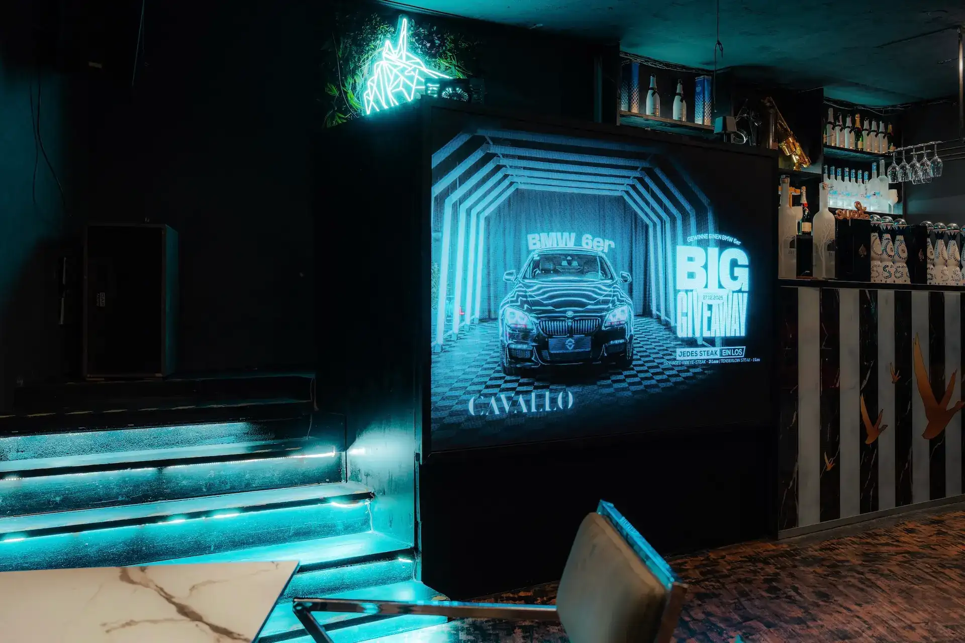 In einem dunklen Raum mit Neonbeleuchtung ist ein großer Bildschirm zu sehen, auf dem eine BMW-Werbung für ein GROSSES GEWINNSPIEL läuft. Auch Regale mit Flaschen und ein Marmortisch sind zu sehen.