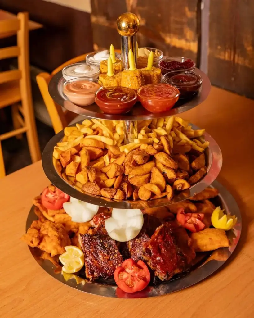 Ein dreistufiges Serviertablett im Centimeter Wien zeigt oben verschiedene Dip-Saucen, in der Mitte Pommes frites und Kartoffelspalten und unten gegrilltes Fleisch mit Beilagen und Zitronenscheiben, alles schön auf einem Holztisch angerichtet.