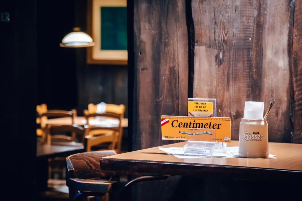 Auf einem Holztisch in einem rustikalen Restaurant in Centimeter Wien stehen eine Centimeter-Lebensmittelbox, Servietten, ein Keramikbehälter mit dem Gösser-Logo und Utensilien; im Hintergrund sind leere Stühle und Tische zu sehen.