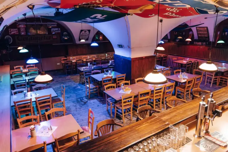 Ein gemütliches, leeres Restaurant mit Holztischen und -stühlen, bunten Hängelampen und dekorativen Schirmen an der Decke. Im Vordergrund ist eine hölzerne Bar mit Gläsern und Bierhähnen zu sehen.