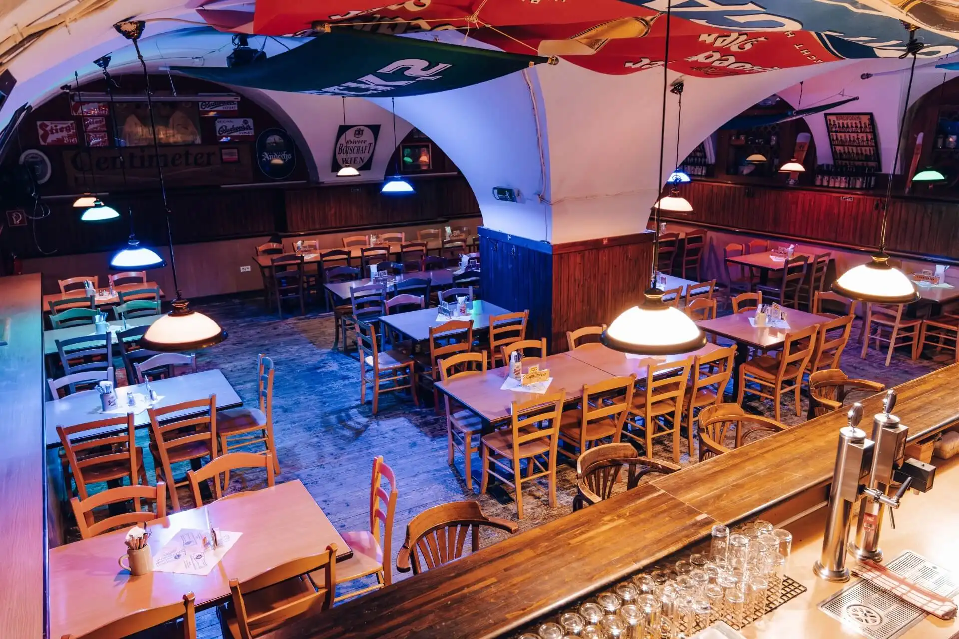 Ein gemütliches, leeres Restaurant mit Holztischen und -stühlen, bunten Hängelampen und einer Bar mit Gläsern und Bierhähnen im Vordergrund. Die Decke ist gewölbt und mit verschiedenen Bannern geschmückt.
