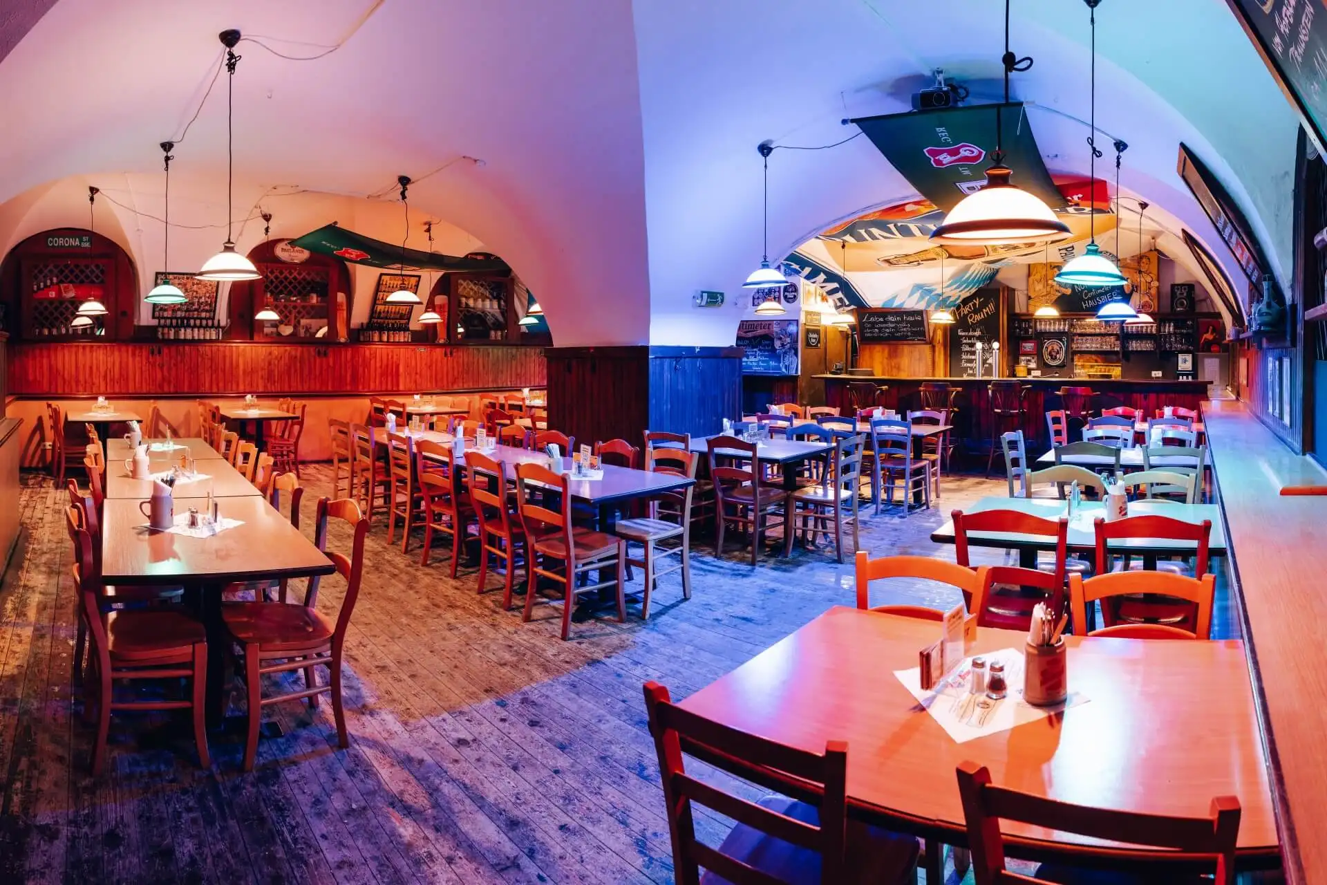 Ein geräumiges, warm beleuchtetes Restaurant mit ordentlich angeordneten Holztischen und -stühlen. Die gewölbte Decke, die Hängelampen und die gemütliche Dekoration schaffen eine einladende Atmosphäre. Das Restaurant scheint leer und bereit für Gäste zu sein.