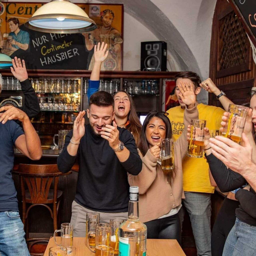 Eine Gruppe fröhlicher Menschen, die Bierkrüge in der Hand halten, hebt die Hände und lächelt, während sie gemeinsam in einer gemütlichen Bar mit Holzdekoration feiern - ein echter Wiener Restaurant-Geheimtipp mit festlicher Atmosphäre.