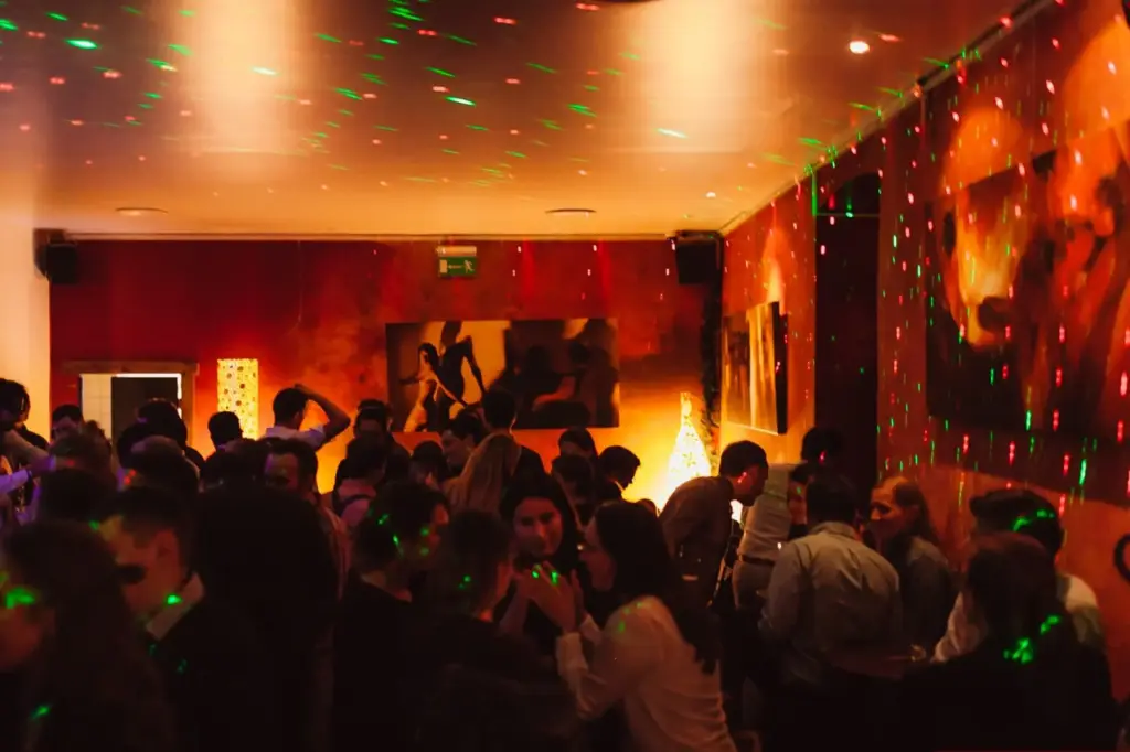 Eine überfüllte Indoor-Party mit Menschen, die bei gedämpftem Licht und bunten grünen Laserlichtern zusammenkommen, mit warmtonigen Wänden, Kunstwerken und beleuchteten Dekorationen im Hintergrund in einer Cocktailbar Wien.