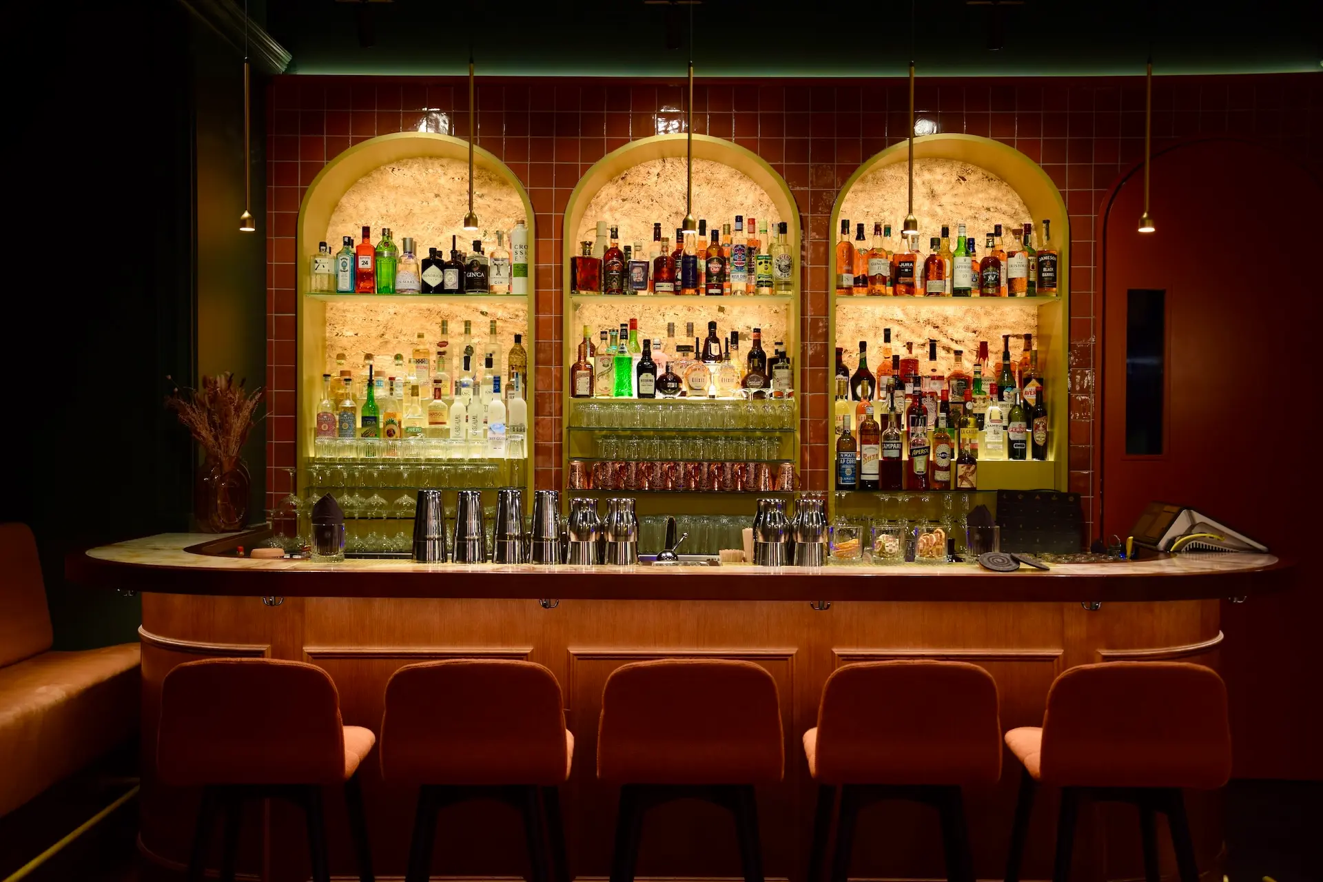 Eine stilvolle Bar mit einer Arbeitsplatte aus Marmor, orangefarbenen gepolsterten Hockern und Regalen mit verschiedenen Spirituosenflaschen, Gläsern und Mixern vor einer warm beleuchteten, gewölbten Kulisse.