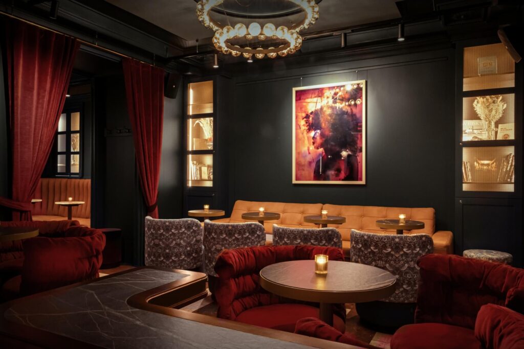 Ein schummriges Wohnzimmer mit roten Samtsesseln, einem braunen Ledersofa, runden Tischen mit Kerzen, dunklen Wänden, goldenen Regalen, Samtvorhängen und einem bunten gerahmten Kunstwerk über dem Sofa.