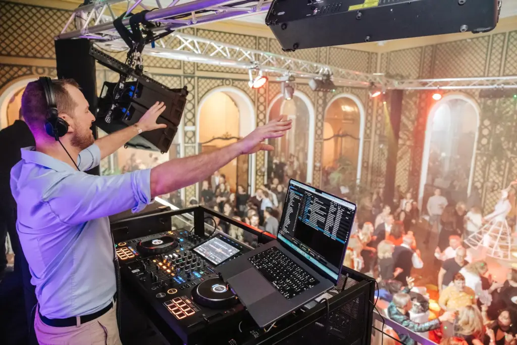 Ein DJ mit Kopfhörern steht an einem Mischpult mit einem Laptop und hebt seine Arme in Richtung einer lebhaften tanzenden Menge in einem geräumigen, verschnörkelten Veranstaltungsort mit bunter Beleuchtung und gewölbten Fenstern.