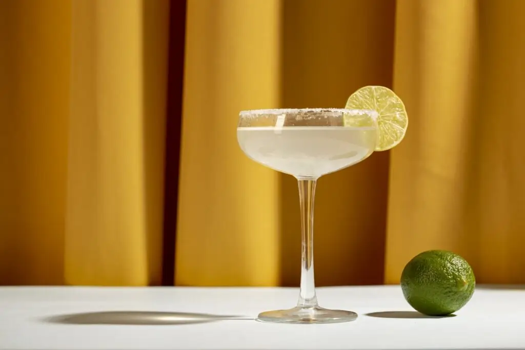 Ein Margarita-Cocktail mit gesalzenem Rand und einem Limettenrad als Garnitur liegt auf einer weißen Fläche neben einer ganzen Limette, mit einem gelben Vorhang im Hintergrund - die perfekte Inspiration für Ihre eigenen Cocktail-Rezepte.