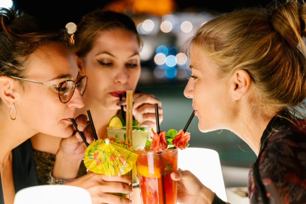 Drei Frauen sitzen nachts in einer Cocktailbar in Wien zusammen, schlürfen bunte Cocktails mit Fruchtgarnituren und Regenschirmen und genießen die Gesellschaft der anderen. Der Hintergrund ist sanft mit Bokeh-Lichtern beleuchtet.