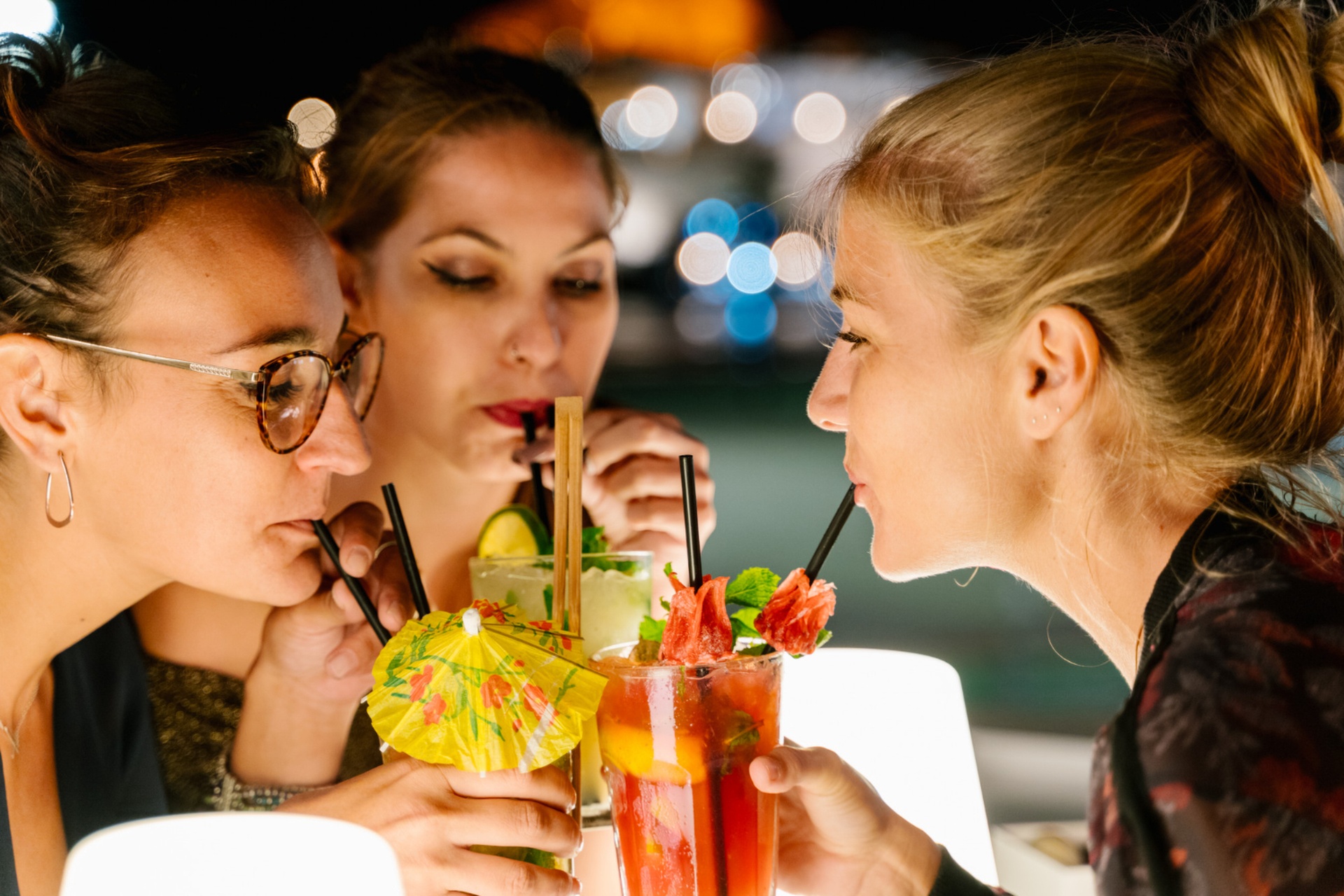 Drei Frauen sitzen nachts in einer Cocktailbar in Wien zusammen, schlürfen bunte Cocktails mit Fruchtgarnituren und Regenschirmen und genießen die Gesellschaft der anderen. Der Hintergrund ist sanft mit Bokeh-Lichtern beleuchtet.