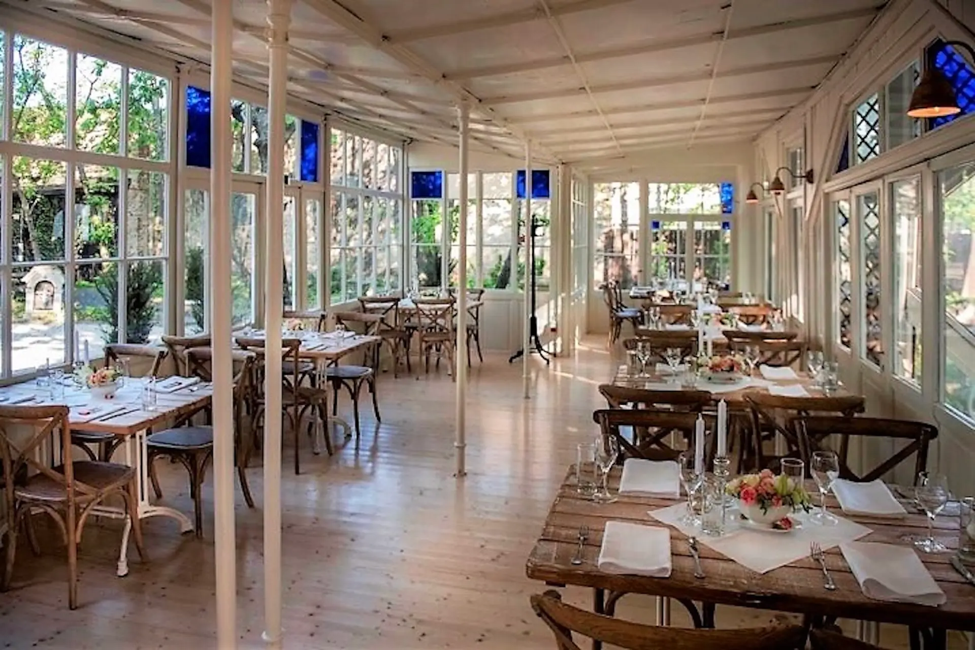 Ein heller, luftiger Restaurantbereich mit Holzböden, weißen Balken, großen Fenstern, rustikalen Holztischen und ordentlich gedeckten Tischen. Das Sonnenlicht strömt herein, und draußen ist das Grün zu sehen.