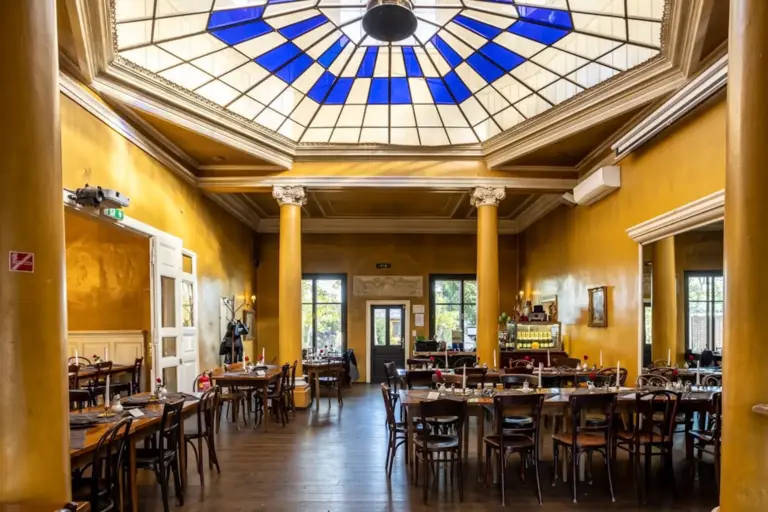 Concordia Schlössl Ein geräumiges Restaurant mit gelben Wänden, großen Säulen, Holztischen und -stühlen und einer blau-weißen, geometrisch gestalteten Glasdecke, die natürliches Licht hereinlässt.