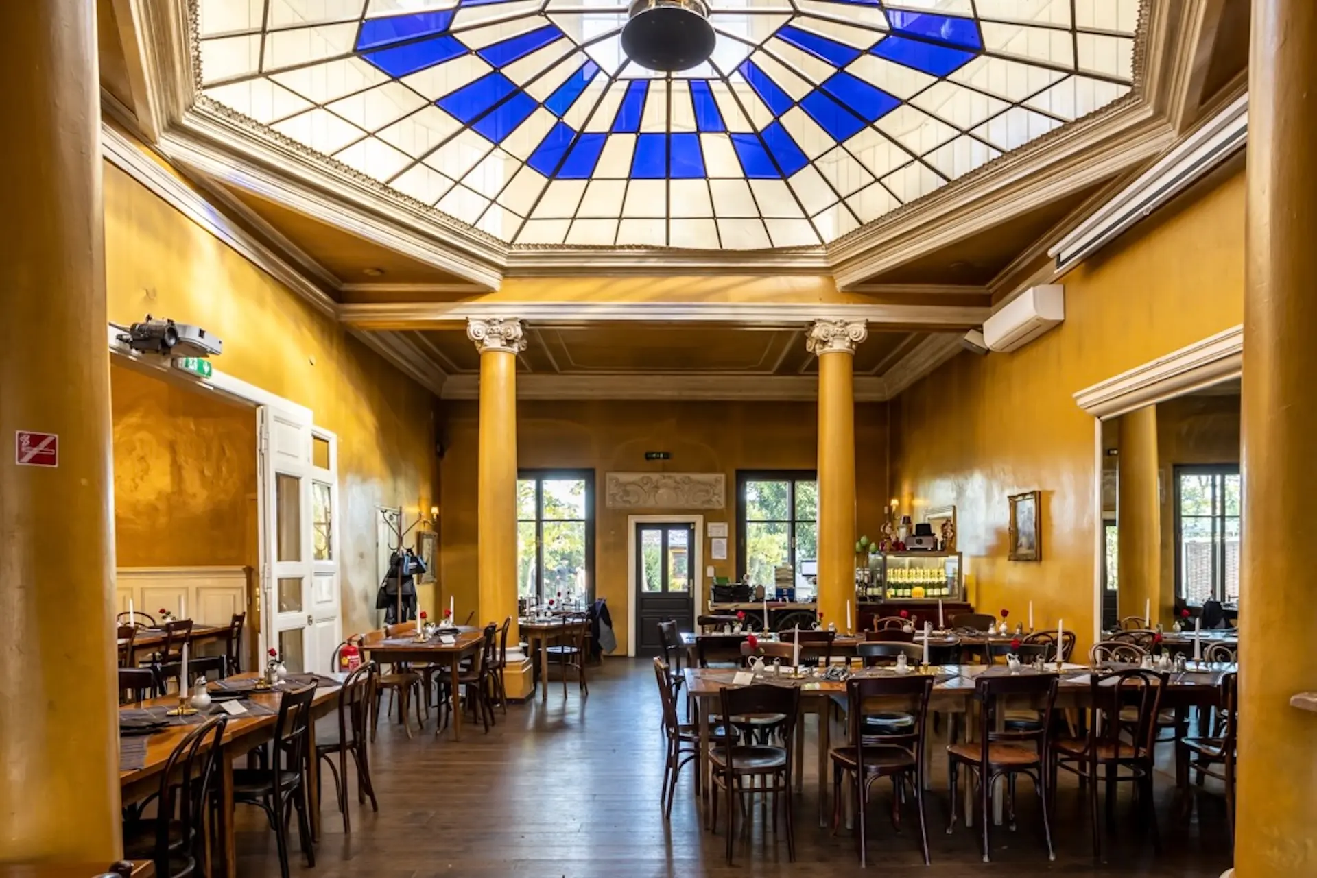 Concordia Schlössl Ein geräumiges Restaurant mit gelben Wänden, großen Säulen, Holztischen und -stühlen und einer blau-weißen, geometrisch gestalteten Glasdecke, die natürliches Licht hereinlässt.