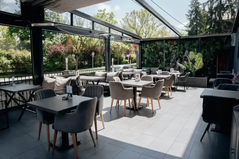 Eine moderne Restaurantterrasse mit grauen gepolsterten Stühlen, dunklen Tischen und Topfpflanzen, umgeben von Glaswänden und Grünpflanzen, unter einem teilweise offenen Dach an einem sonnigen Tag.