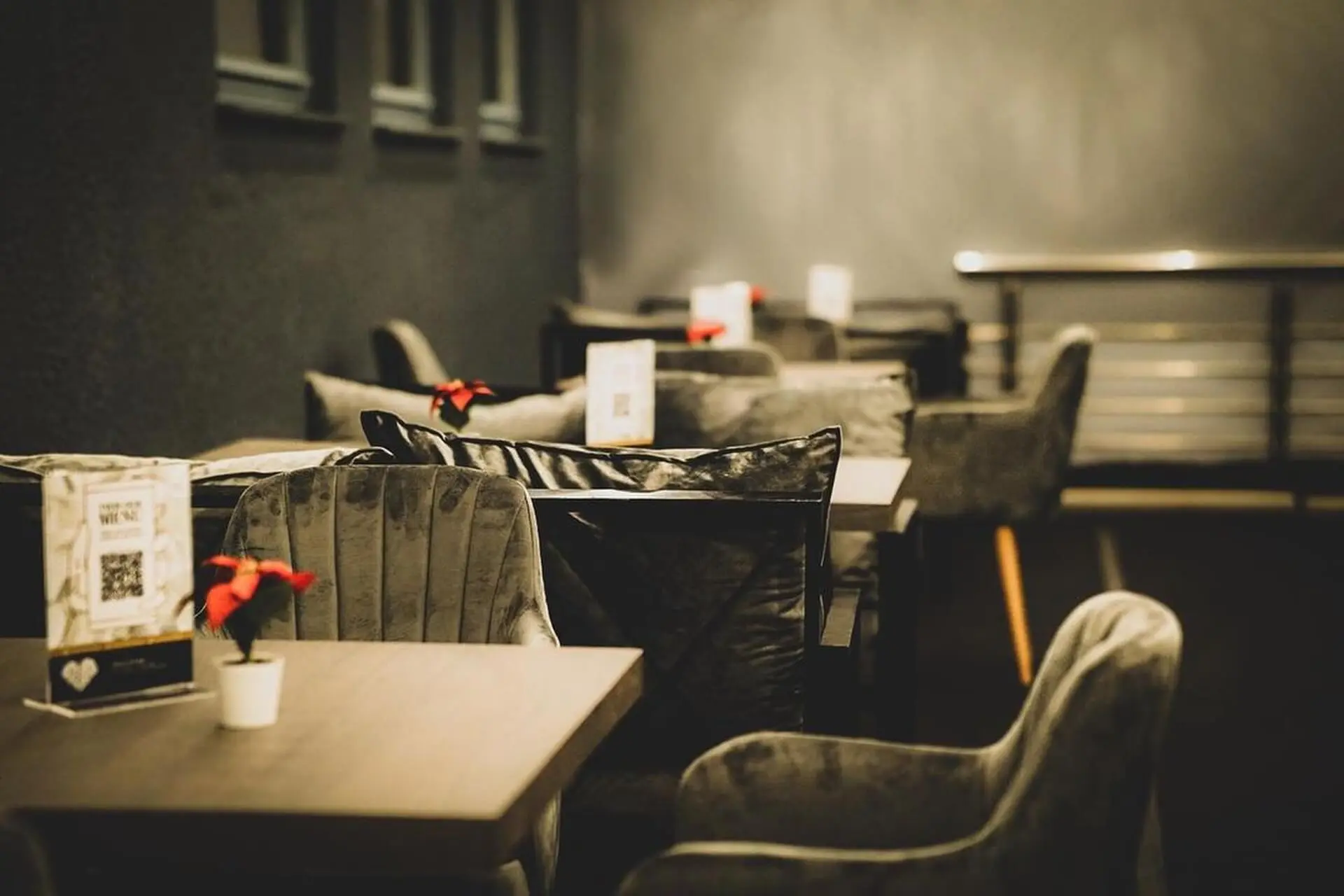 Ein modernes, schummriges Café mit grauen Plüschsesseln, kleinen Holztischen und roten Topfblumen in der Mitte. Auf jedem Tisch sind Speisekarten und ein QR-Code für die Bestellung angebracht.