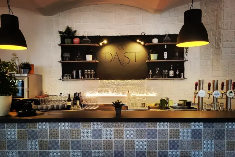 Eine moderne Bar mit gemusterter Fliesenfront, Regalen mit Gläsern und Flaschen, zwei großen Pendelleuchten und einem schwarzen Kreideschild mit der Aufschrift DAST auf einer weißen, strukturierten Wand.